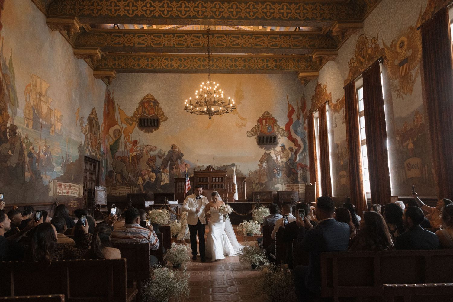 Elegant wedding ceremony in ornate hall with murals, chandelier, and guests seated in pews.