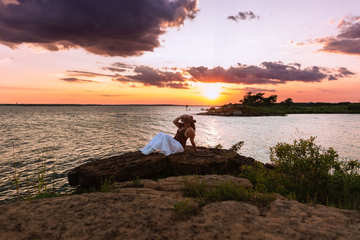 Couple embracing on rocky shoreline at golden sunset with dramatic clouds over calm water.