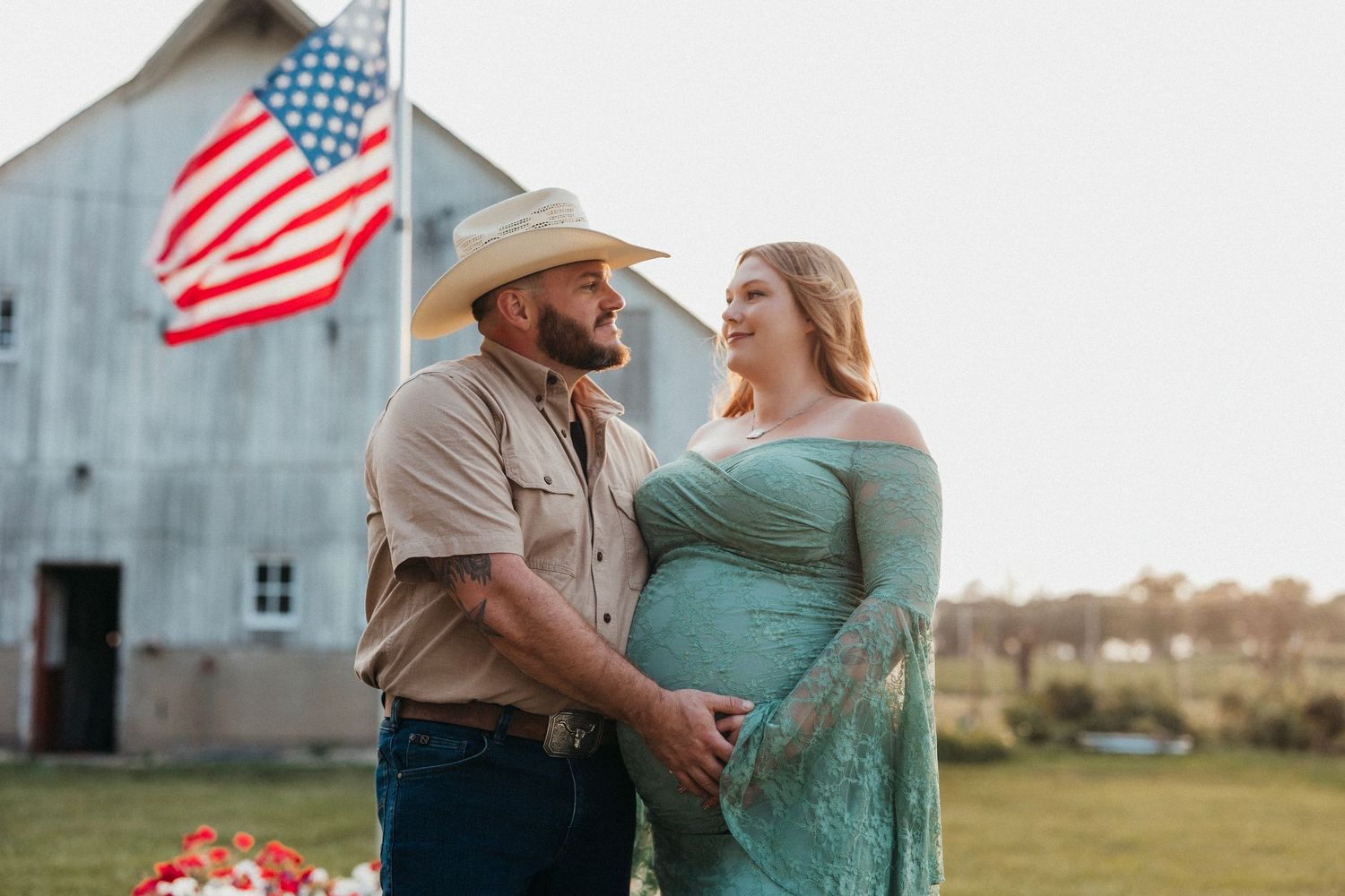 Pregnant couple poses lovingly before rustic barn with American flag in soft evening light.