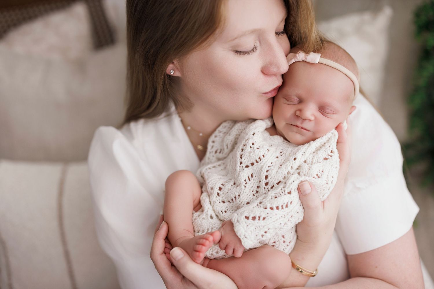 A mother tenderly kisses her sleeping newborn baby wrapped in a white lace swaddle with a floral headband.