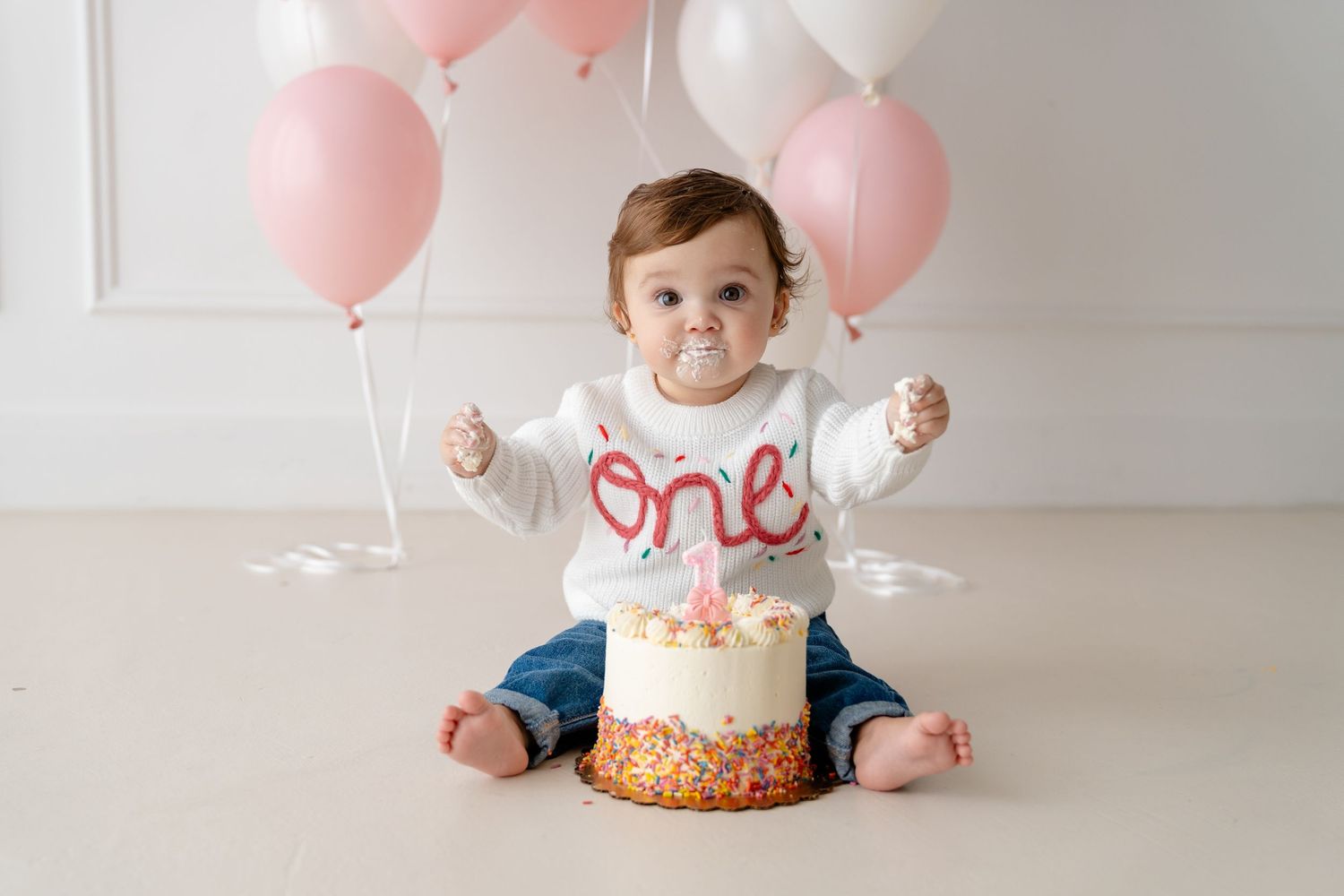 Baby in a white "one" sweater sits before a smash cake surrounded by pink and white balloons on their first birthday.