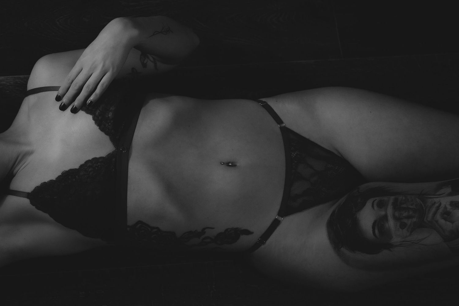 Zwart wit boudoir foto van een vrouw liggend met billen omhoog in een donker lingerie setje met een raam op de achergrond en zonnestrallen