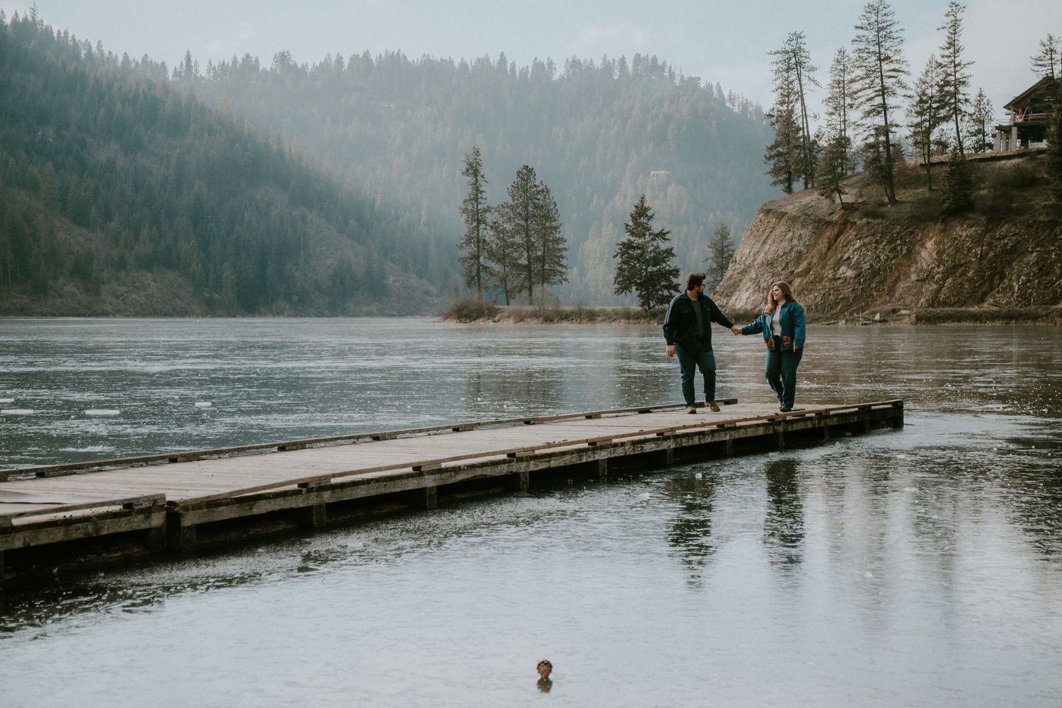 Two people sitting on a wooden dock by a lake surrounded by trees and rocky cliffs.