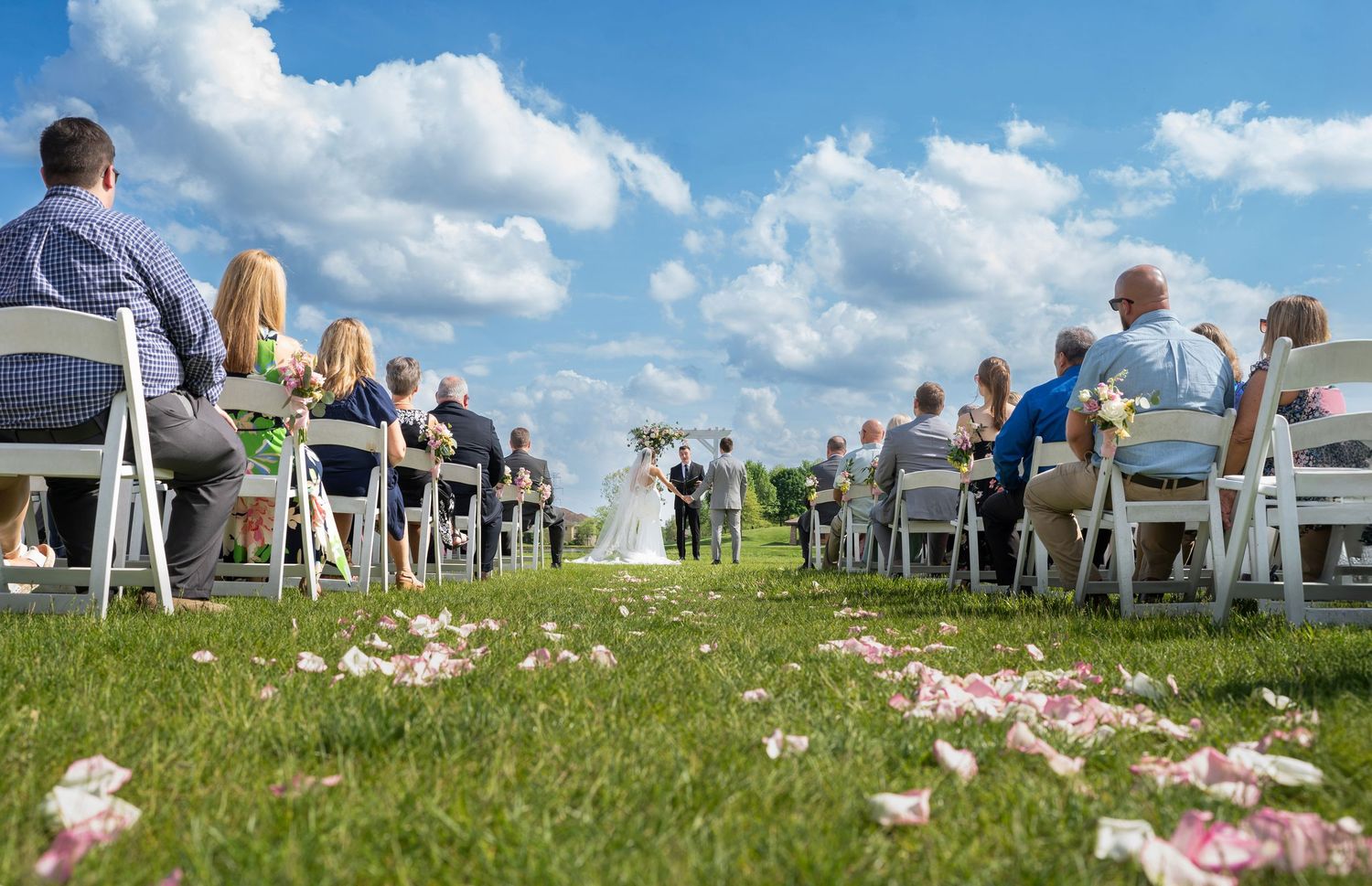 Outdoor wedding ceremony on lush green grass with guests seated in white chairs under a bright blue sky with fluffy clouds.
