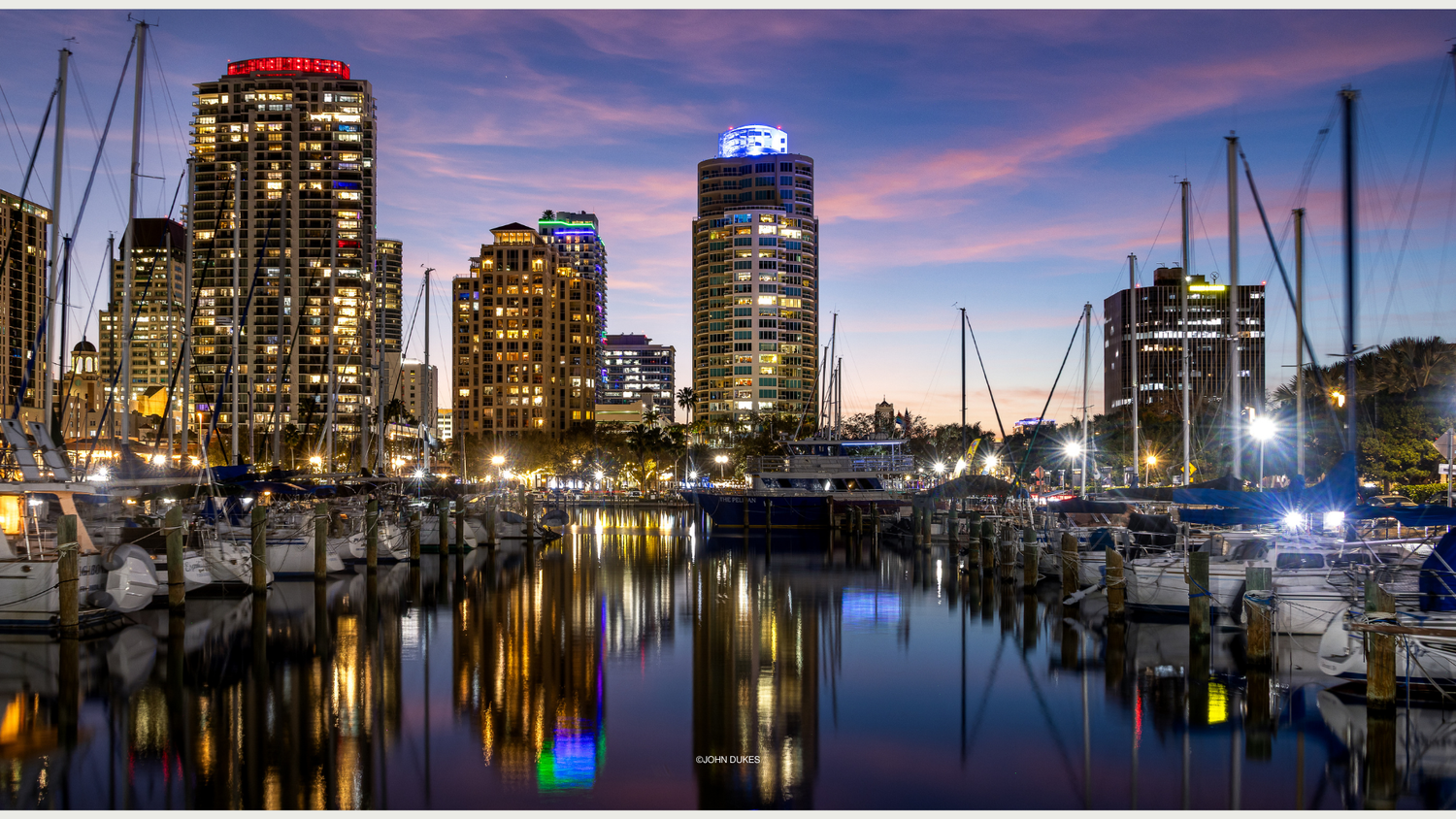 City marina at dusk with glowing skyline reflections on calm water, sailboats docked under a vibrant purple and pink sunset sky.
