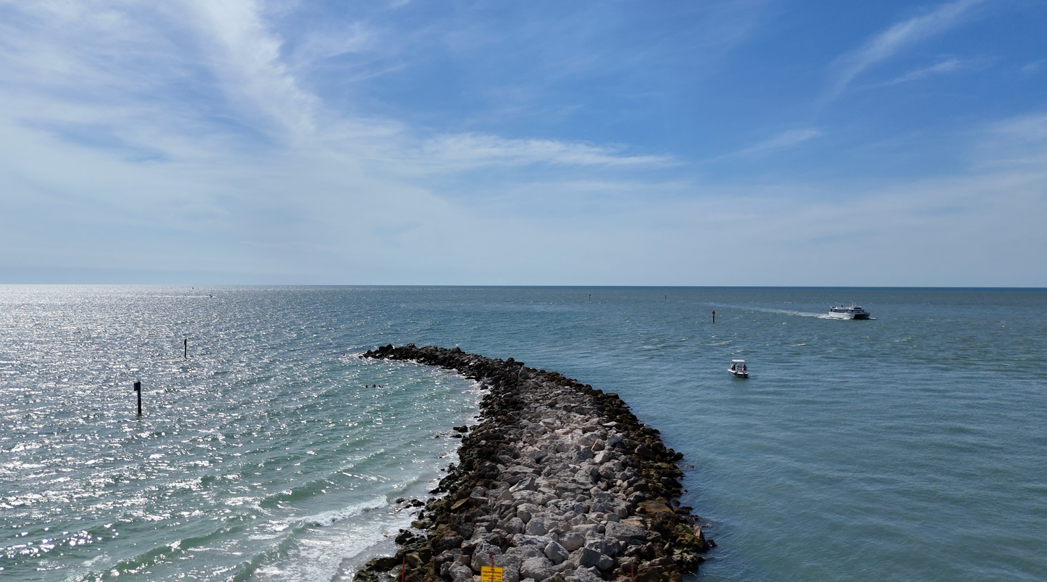 Aerial view of a rocky jetty extending into calm turquoise ocean waters under a bright blue sky with wispy clouds.