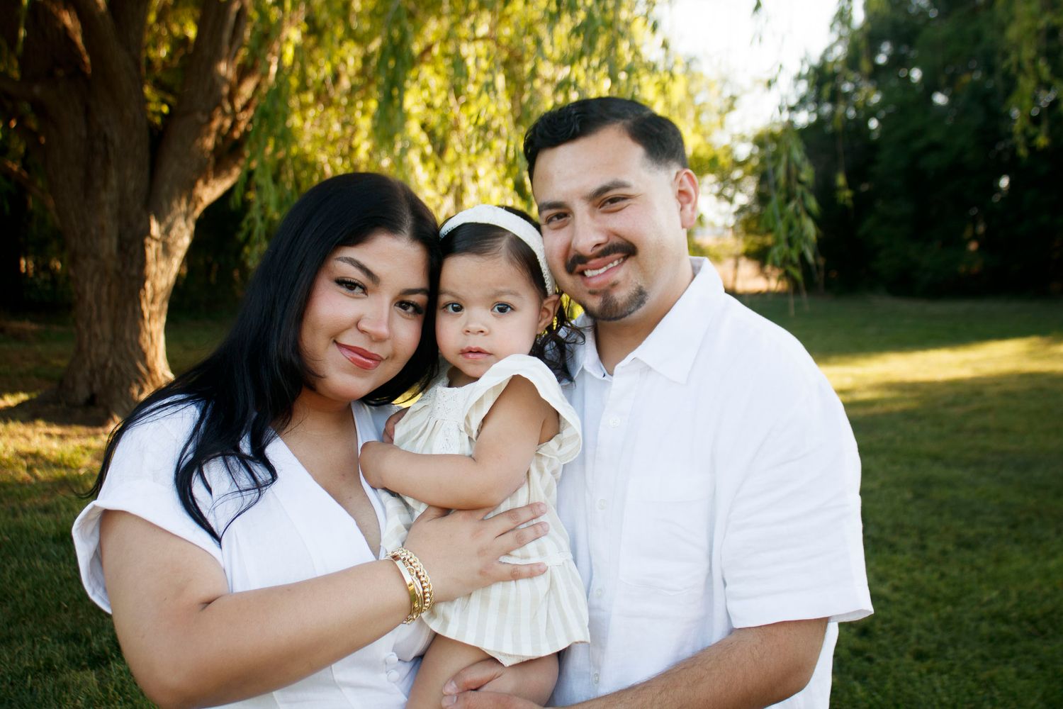 Happy Hispanic family with parents holding their toddler daughter outdoors in a lush green park setting in Canyon, Texas