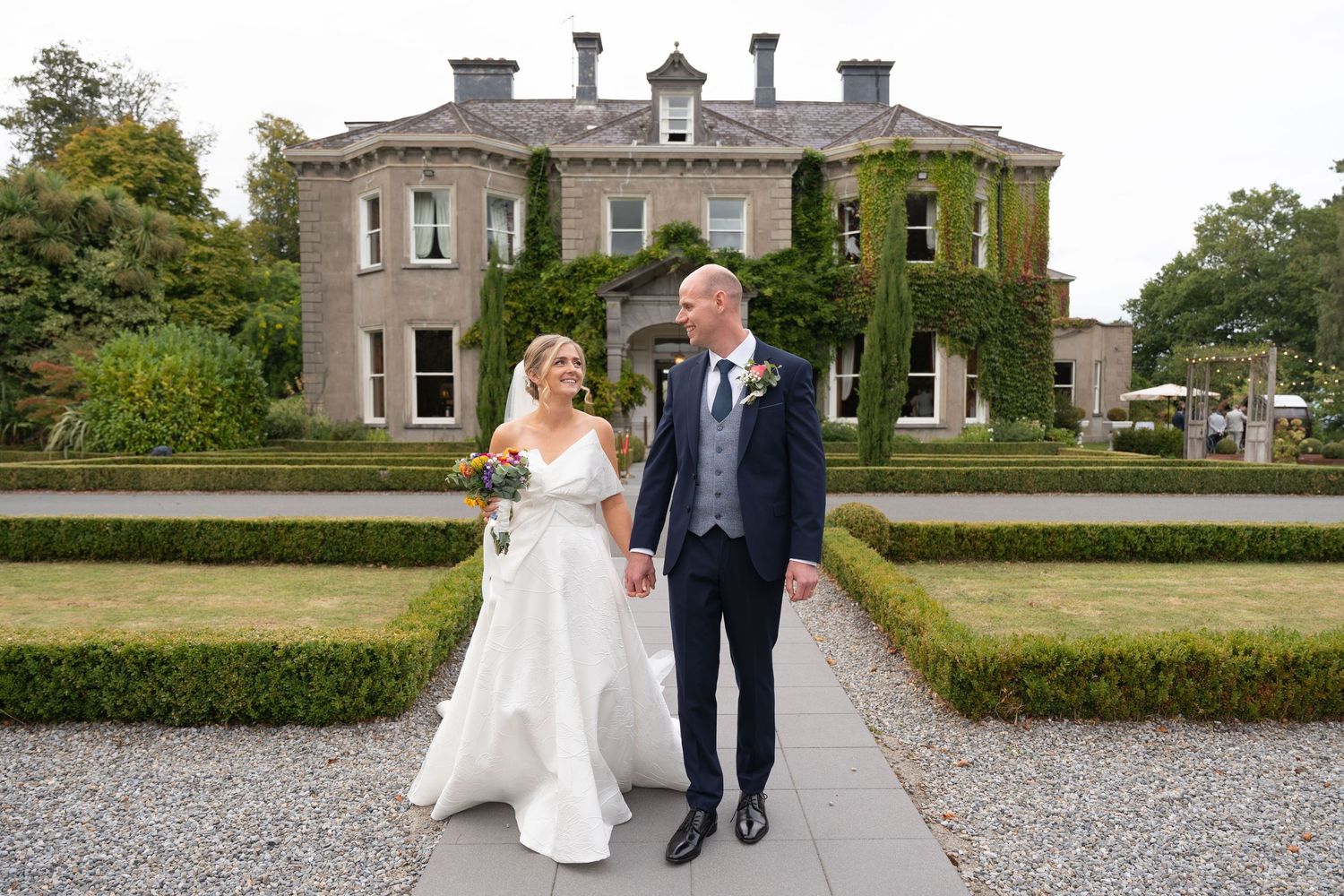 Bride and groom holding hands walking in formal gardens outside an elegant ivy-covered manor house wedding venue.