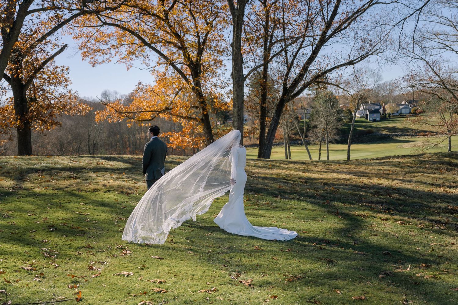 Bride and groom walk through autumn park, long veil flowing in wind among colorful fall trees on green lawn.