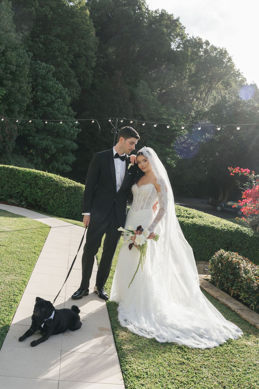 Bride in white gown and groom in tuxedo pose with black dog on garden path surrounded by lush greenery.