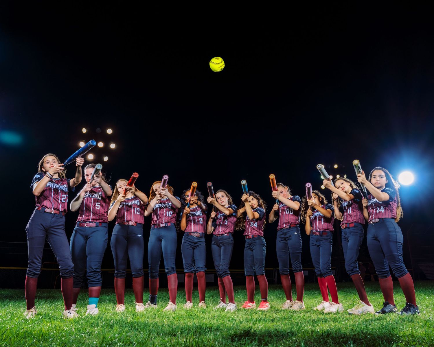 Softball team posing with bats raised under bright stadium lights, yellow softball visible in night sky.