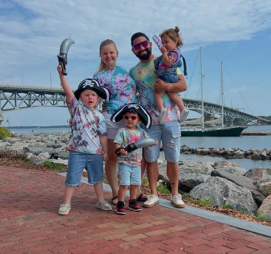 A cheerful family of five in tie-dye shirts poses near a harbor with a sailboat.