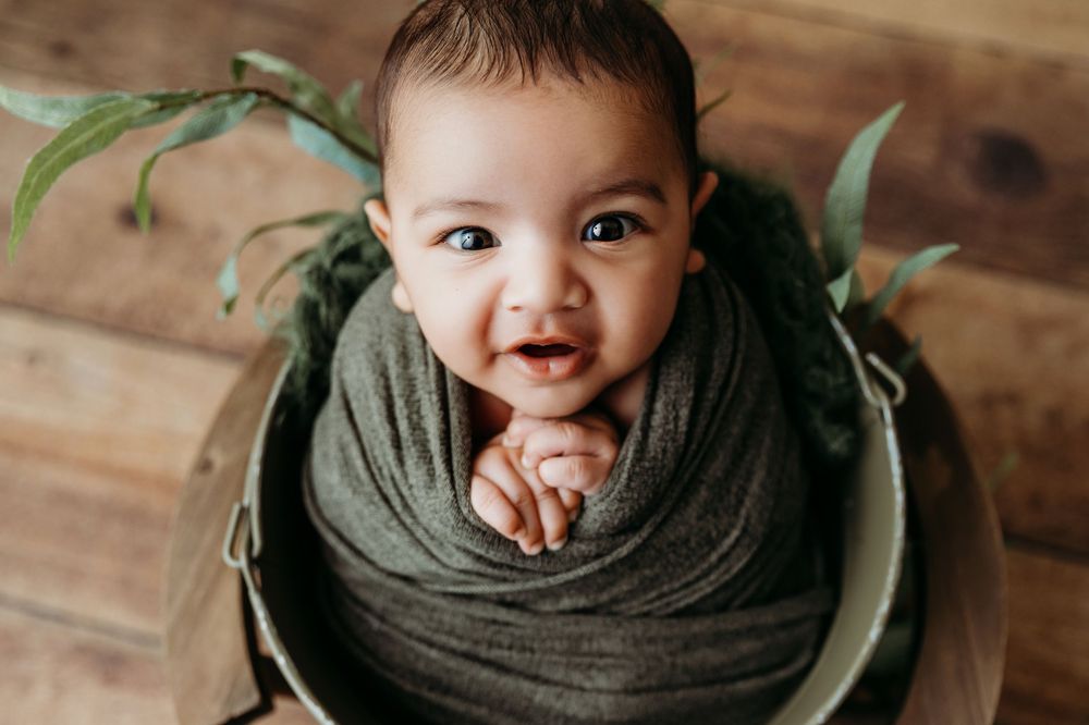 A baby wearing an olive green sweater smiles and plays while lying on wooden floor panels.