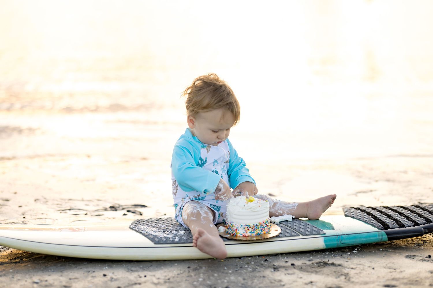 Baby sitting on a surfboard at the beach, wearing a teal outfit, playing with a small cake in golden sunlight.