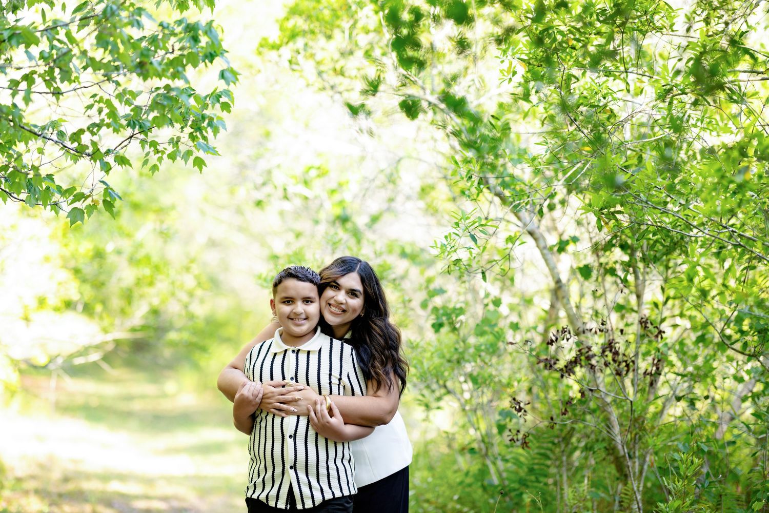 A woman and child embrace on a tree-lined path surrounded by lush green foliage outdoors.
