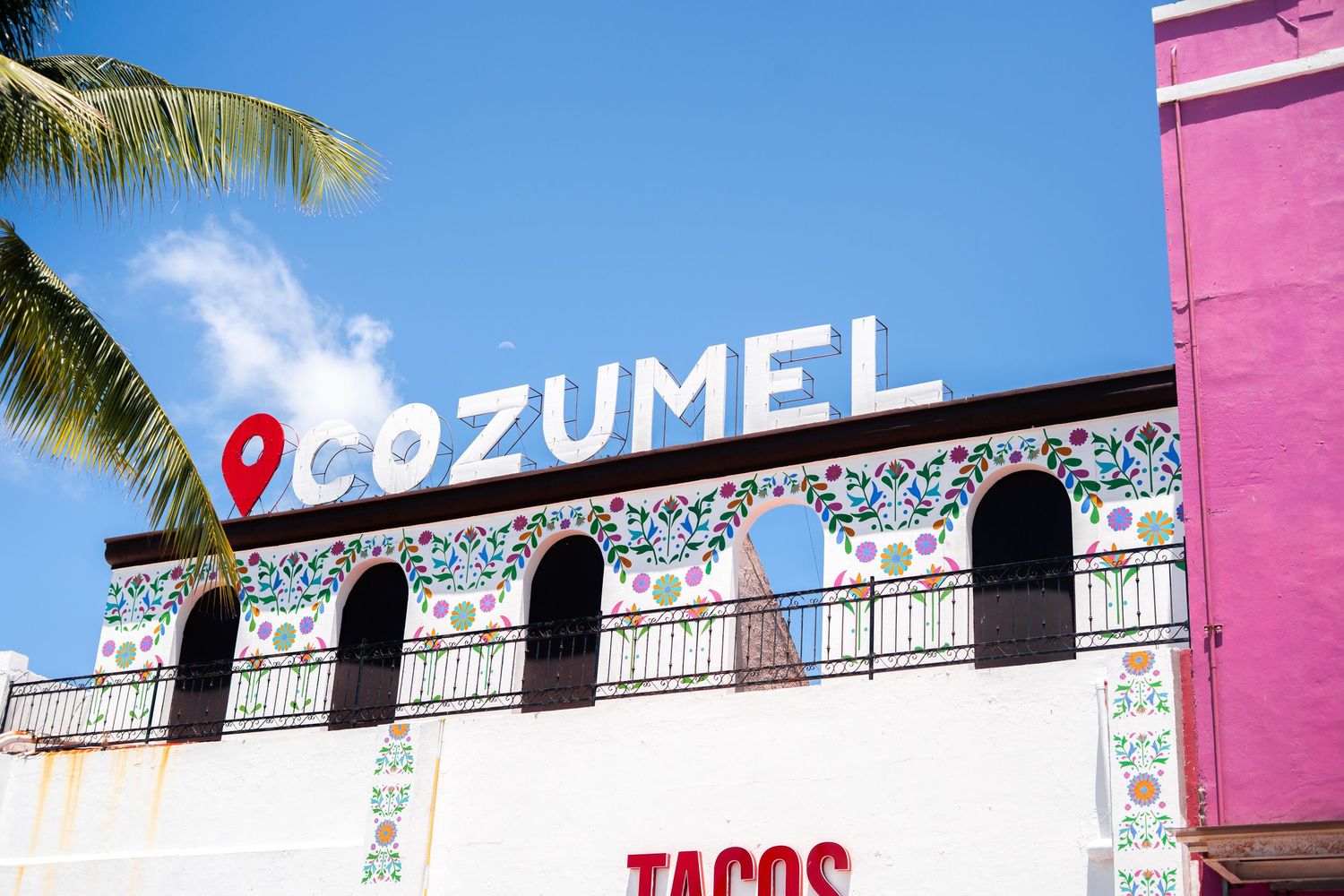 Mexican flag waving against blue sky with palm tree and thatched roof structure in tropical setting.