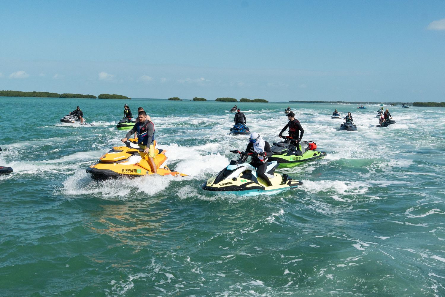 Group of jet skiers riding across blue ocean waters near a tropical coastline.