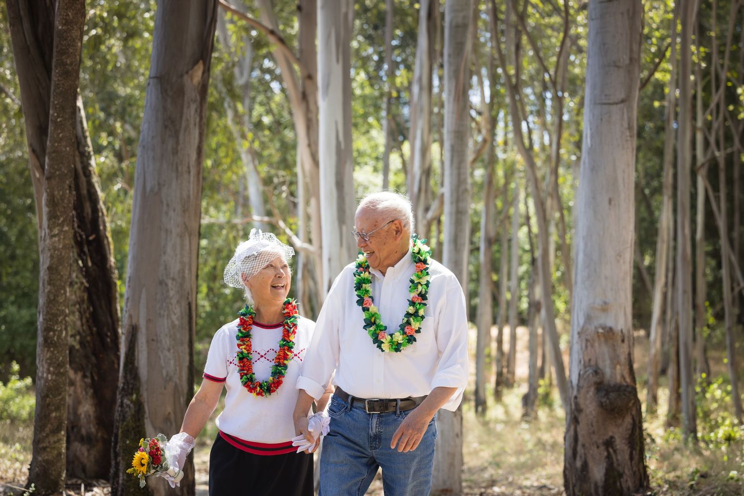 Elderly couple wearing colorful leis walk hand in hand through a sunlit eucalyptus forest.