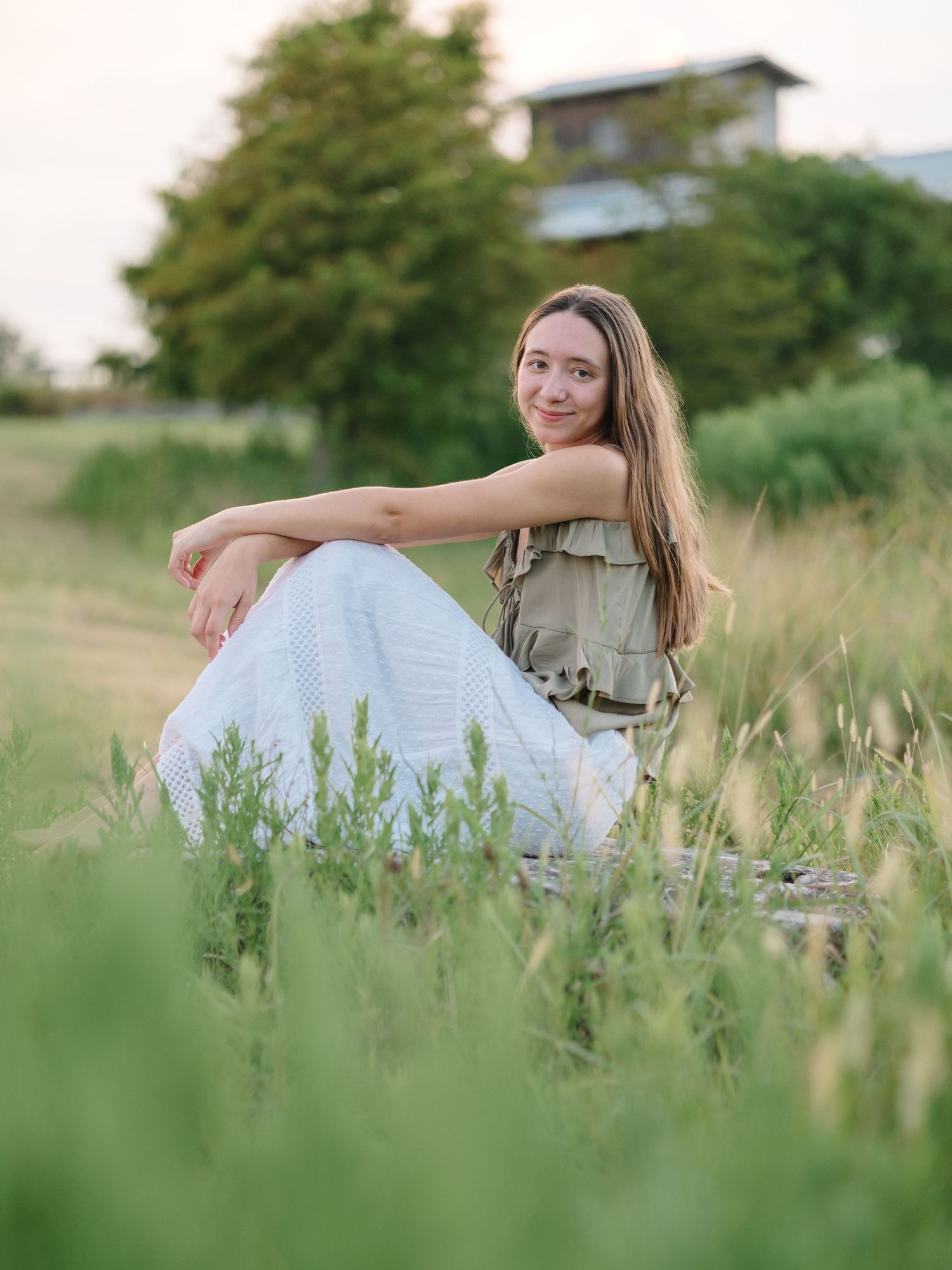 Young woman in white pants and olive top sitting in lush green grass field, smiling outdoors at golden hour.