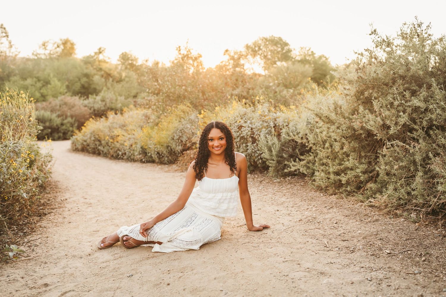 A young woman in a blue dress and cowboy boots stands smiling in a natural outdoor setting with trees and rocks.