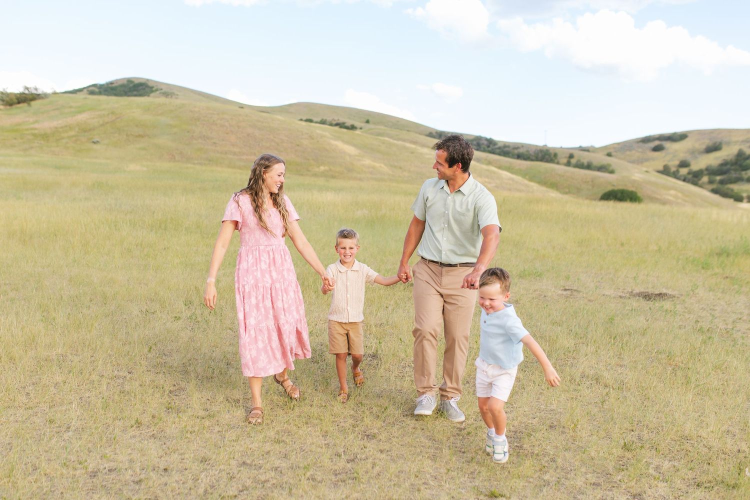 Family of four walking hand-in-hand through green rolling hills on a sunny day.
