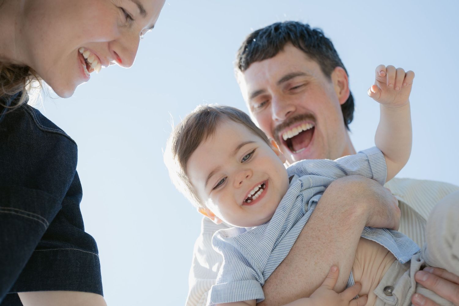 Low-angle shot of a laughing family of three against a bright West Vancouver sky. Joyful, candid family portraits by Tash Takes Pics, North
