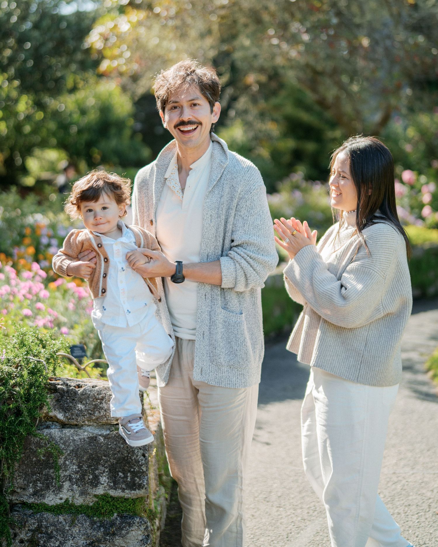 Happy family with mustached dad holding toddler and smiling mom in a sunny garden setting.
