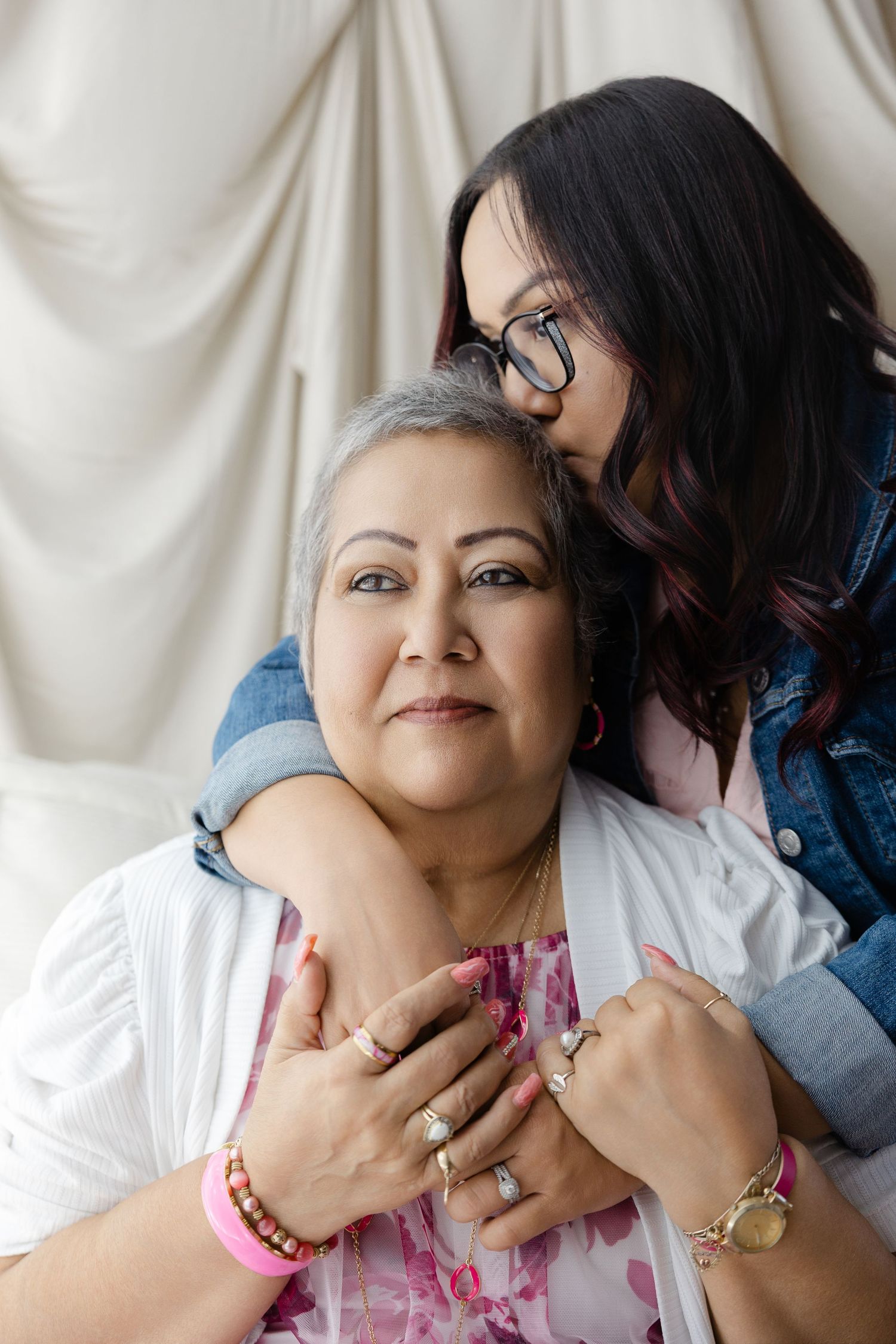A younger woman hugs an older woman from behind, both smiling warmly in a tender mother-daughter portrait.