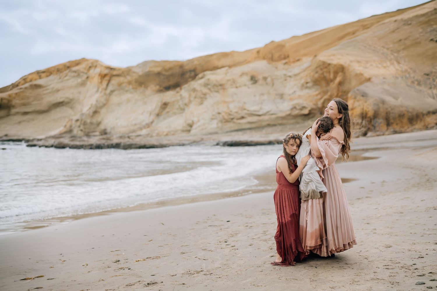 A mother and daughter in boho dresses pose on a sandy beach with ocean waves and golden cliffs behind them.
