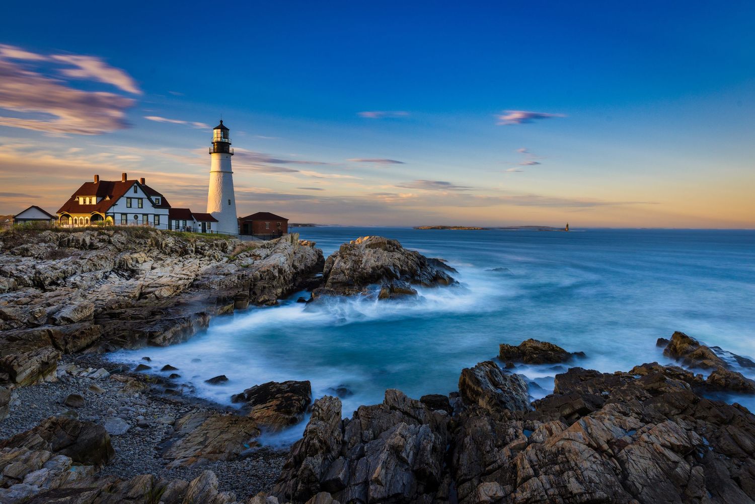White lighthouse on rocky Maine coast at sunset with blue sky and misty ocean waves crashing below.