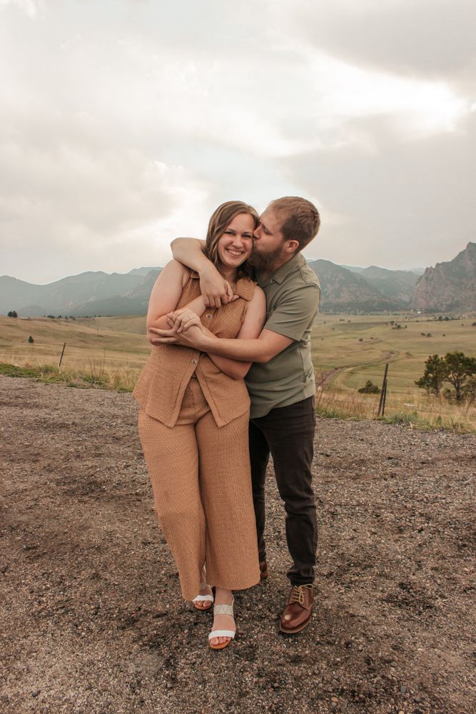 Couple embracing outdoors as man kisses her cheek with stunning mountain landscape backdrop | What to Wear for Family Photos