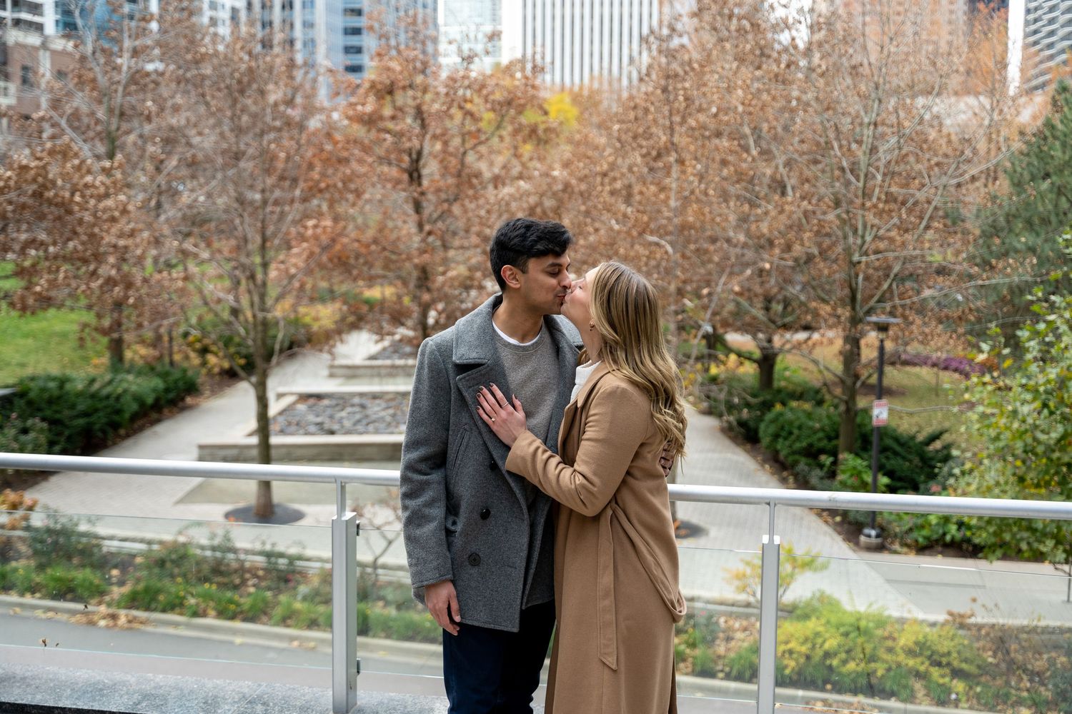 A couple walks together through an autumn park with tall city buildings visible in the background.