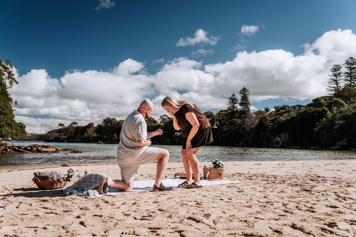 Man proposing to woman on sandy beach with picnic basket, blue sky, and lush trees in background.