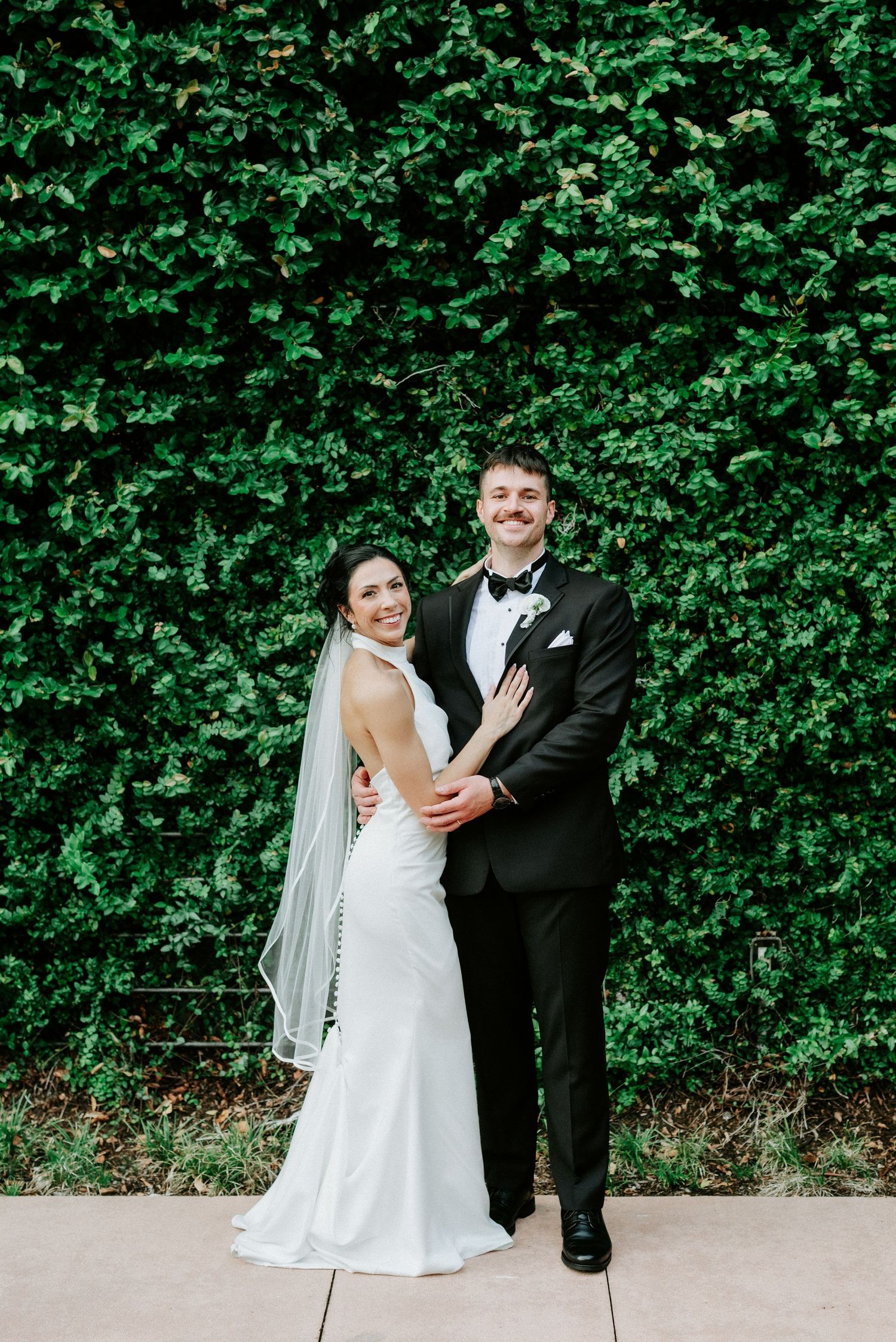 A smiling bride in a white gown and veil poses with a groom in a black tuxedo before a lush green ivy wall.