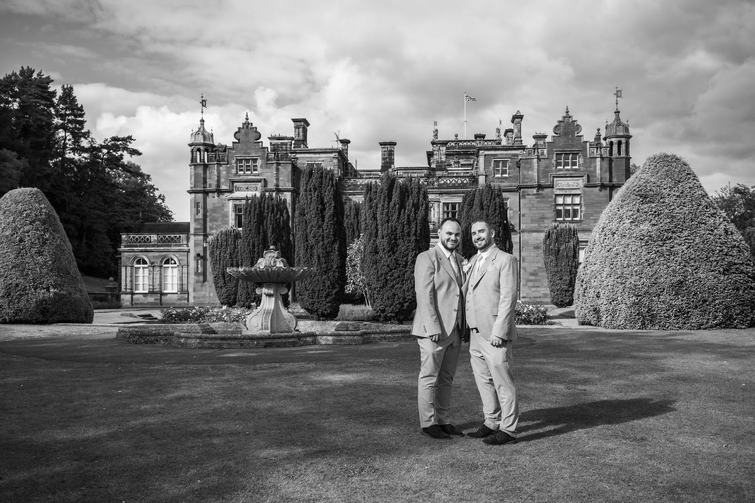 Two men in suits pose together on a lawn before a grand historic manor house in this elegant black and white photo.