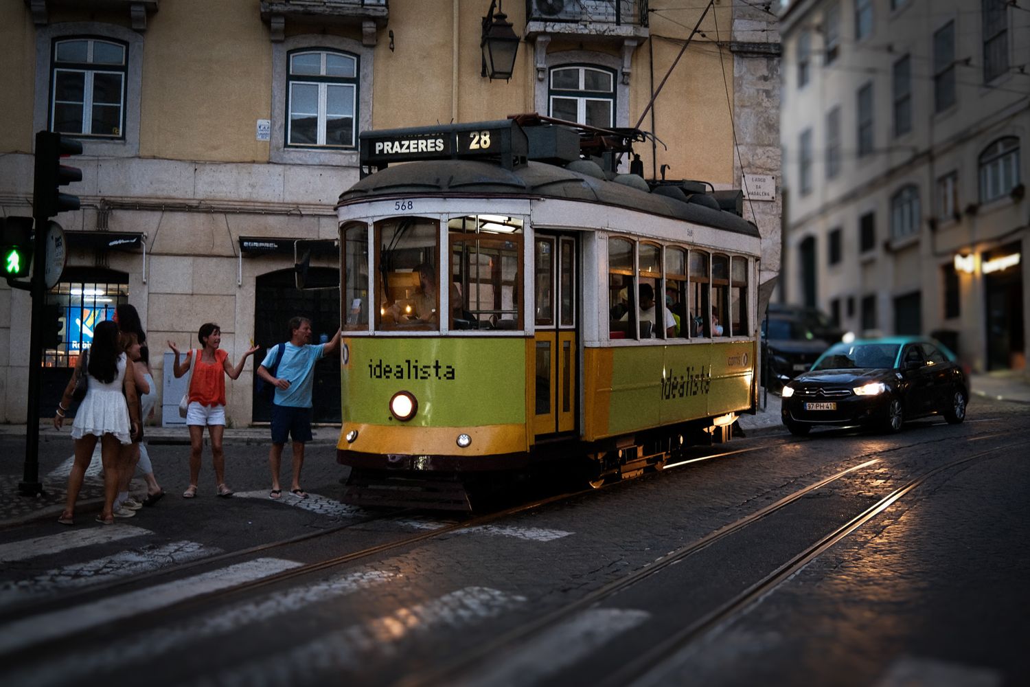 Iconic yellow Lisbon tram number 28 navigates a wet cobblestone street at dusk with pedestrians nearby.