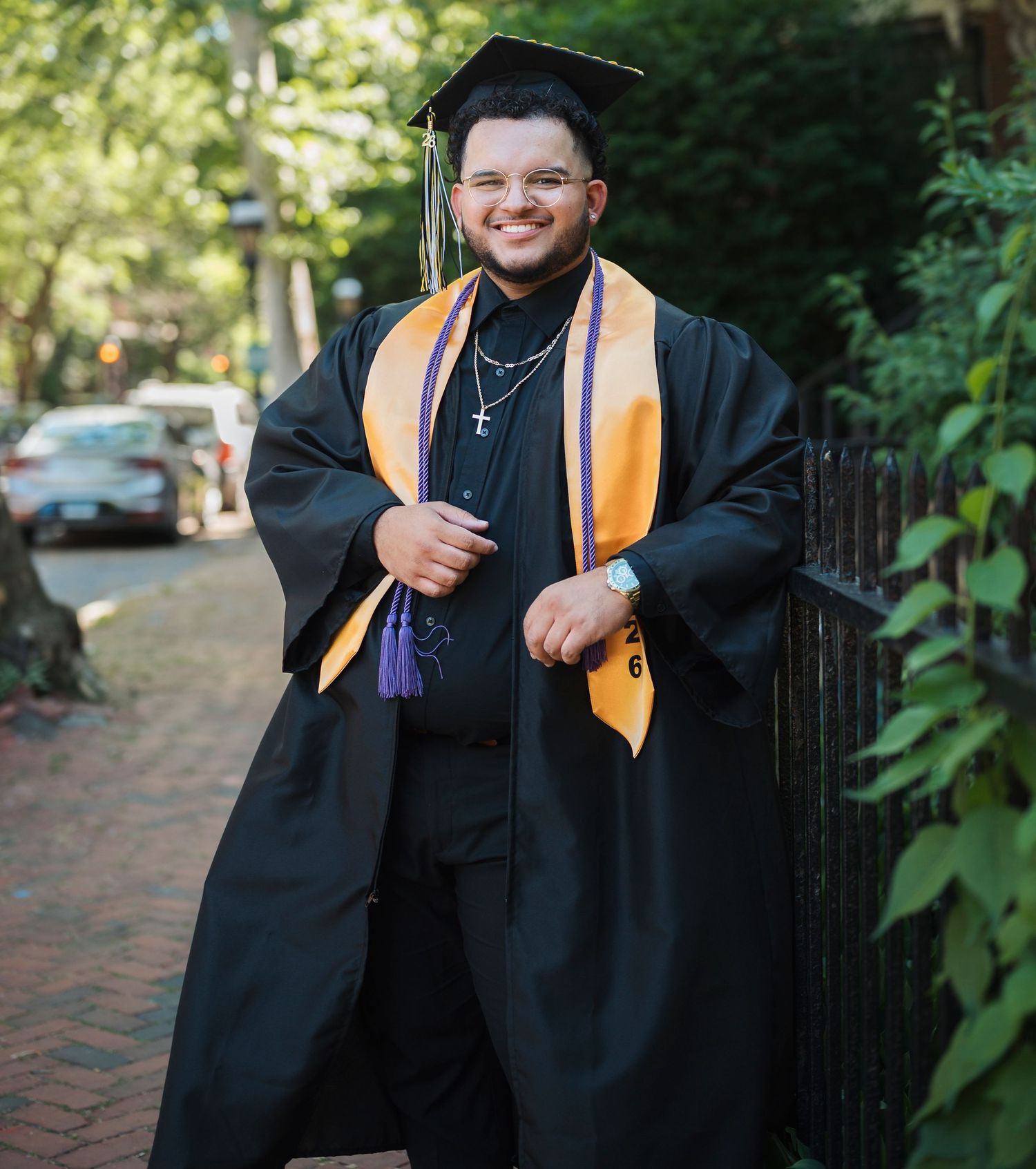 Smiling graduate in black cap and gown with gold and purple stole poses on brick path outdoors.