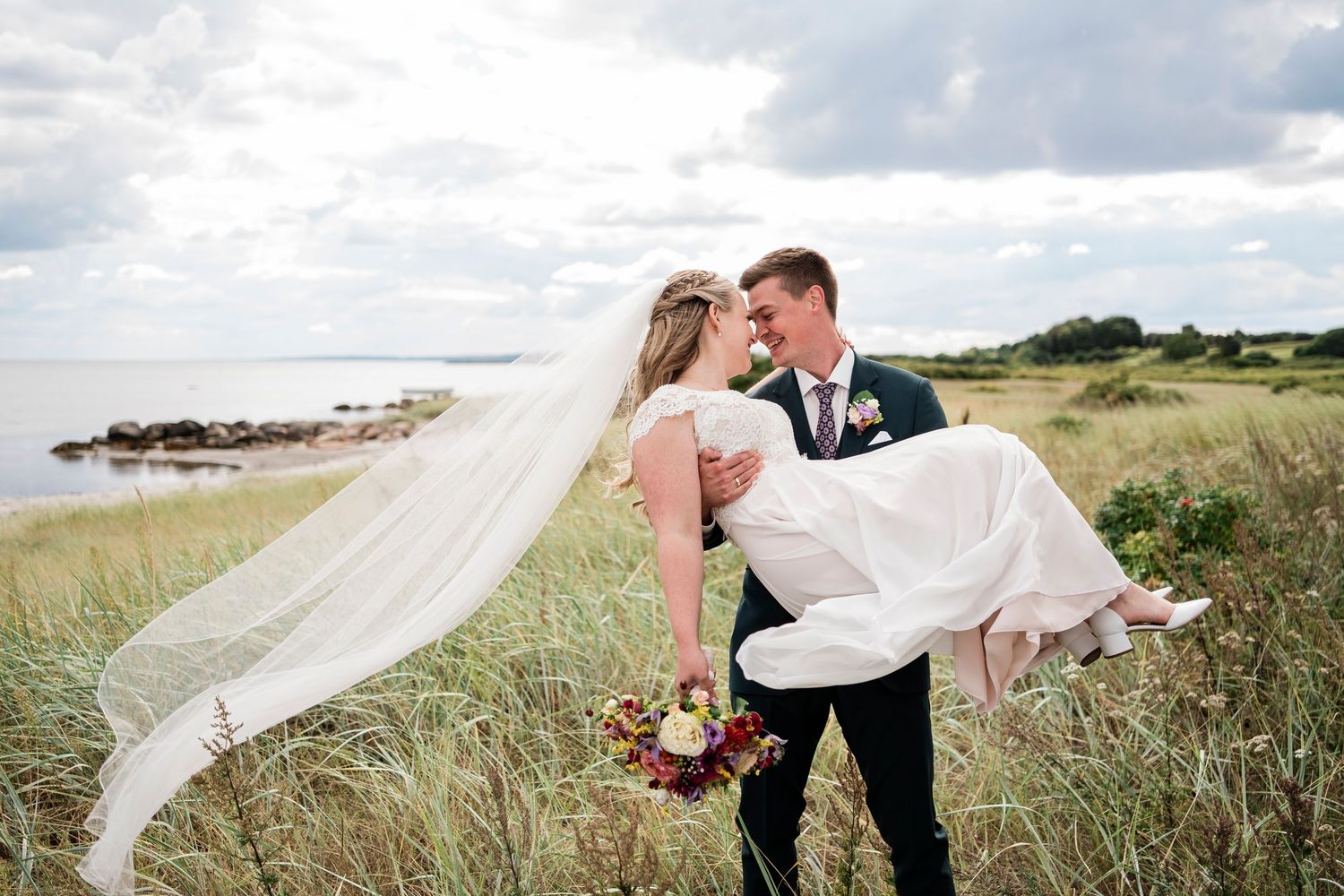 Romantic coastal wedding portrait with couple embracing among beach grass at sunset.