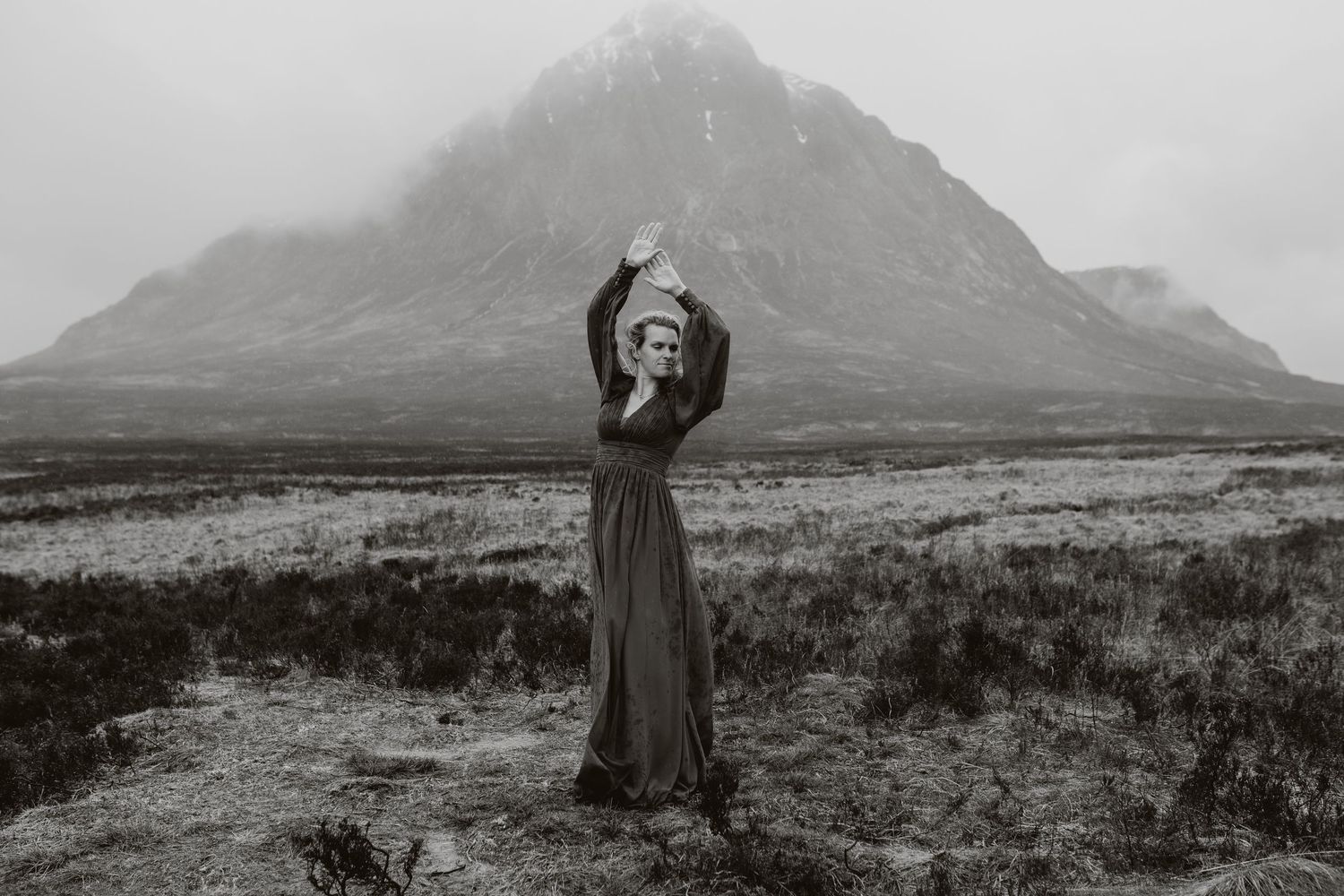Woman in long dark dress poses with arms raised before misty mountain landscape in black and white.