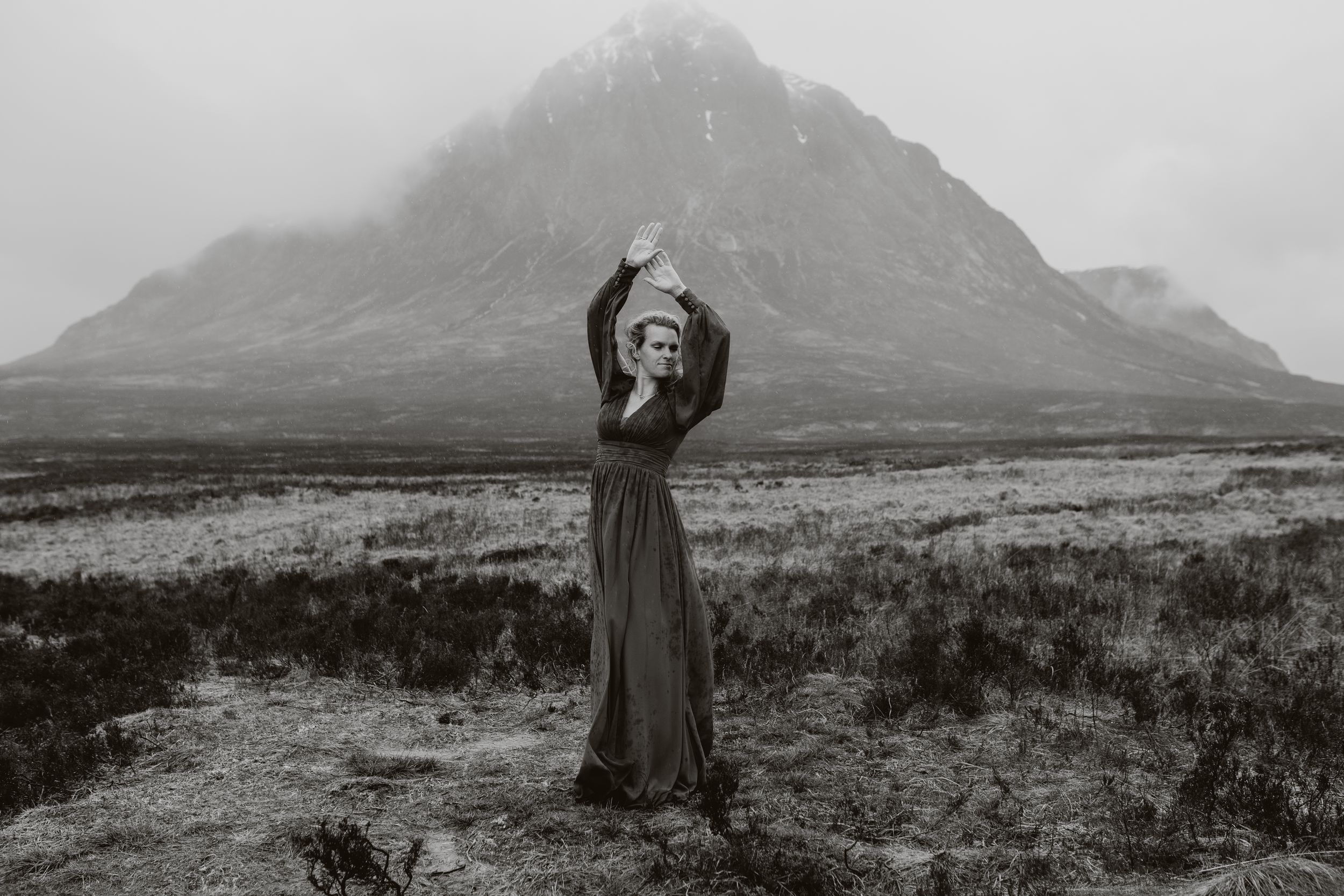 Woman in long dark dress poses with arms raised before misty mountain landscape in black and white.
