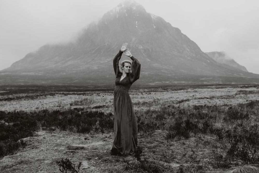 Woman in long dark dress poses with arms raised before misty mountain landscape in black and white.