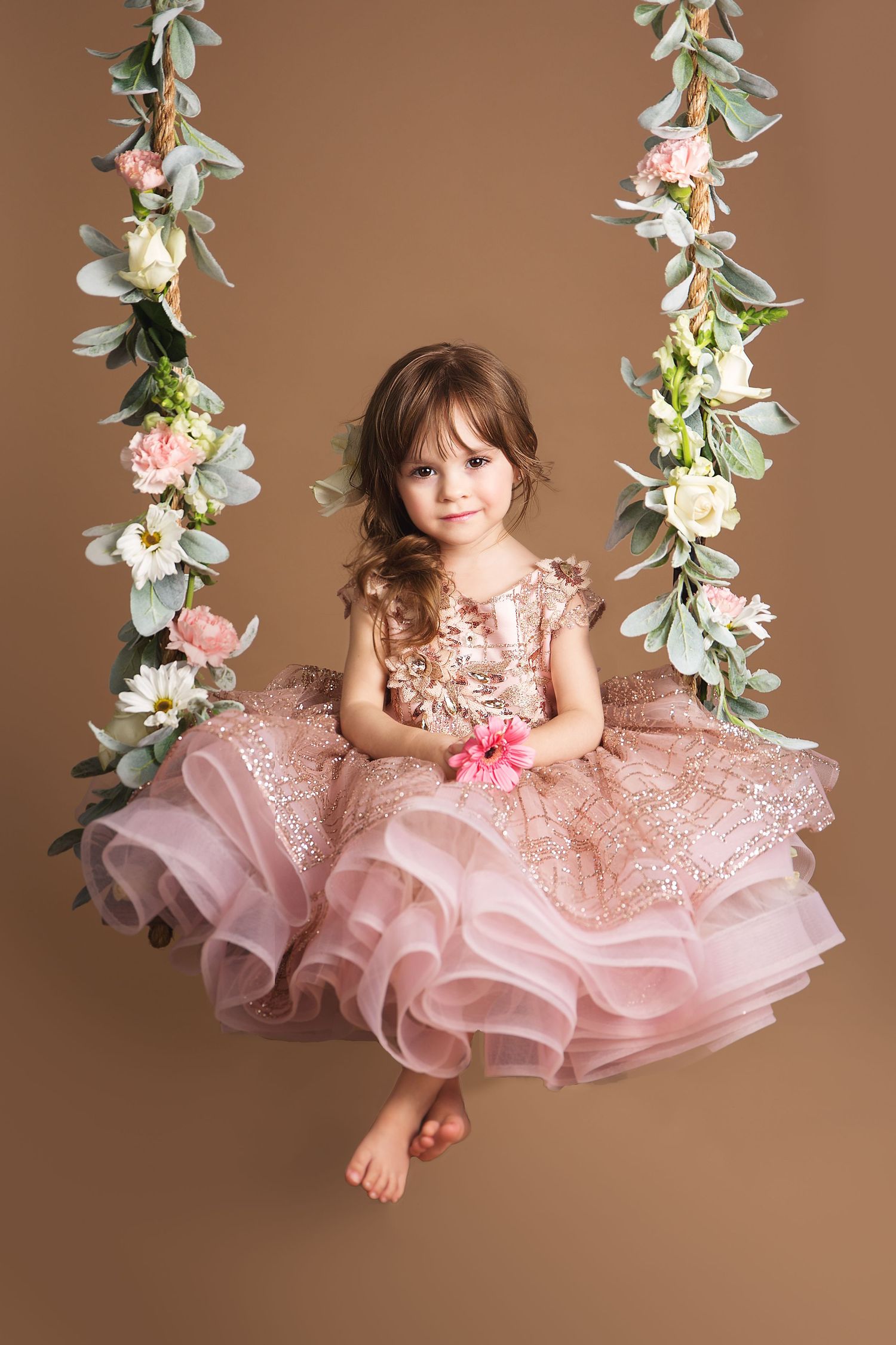 A small child wearing a pink lace dress swings gracefully on a floral swing against a brown backdrop.