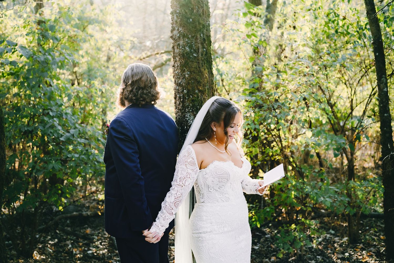 Two people in wedding attire walk together through a sunlit wooded area during an autumn day.