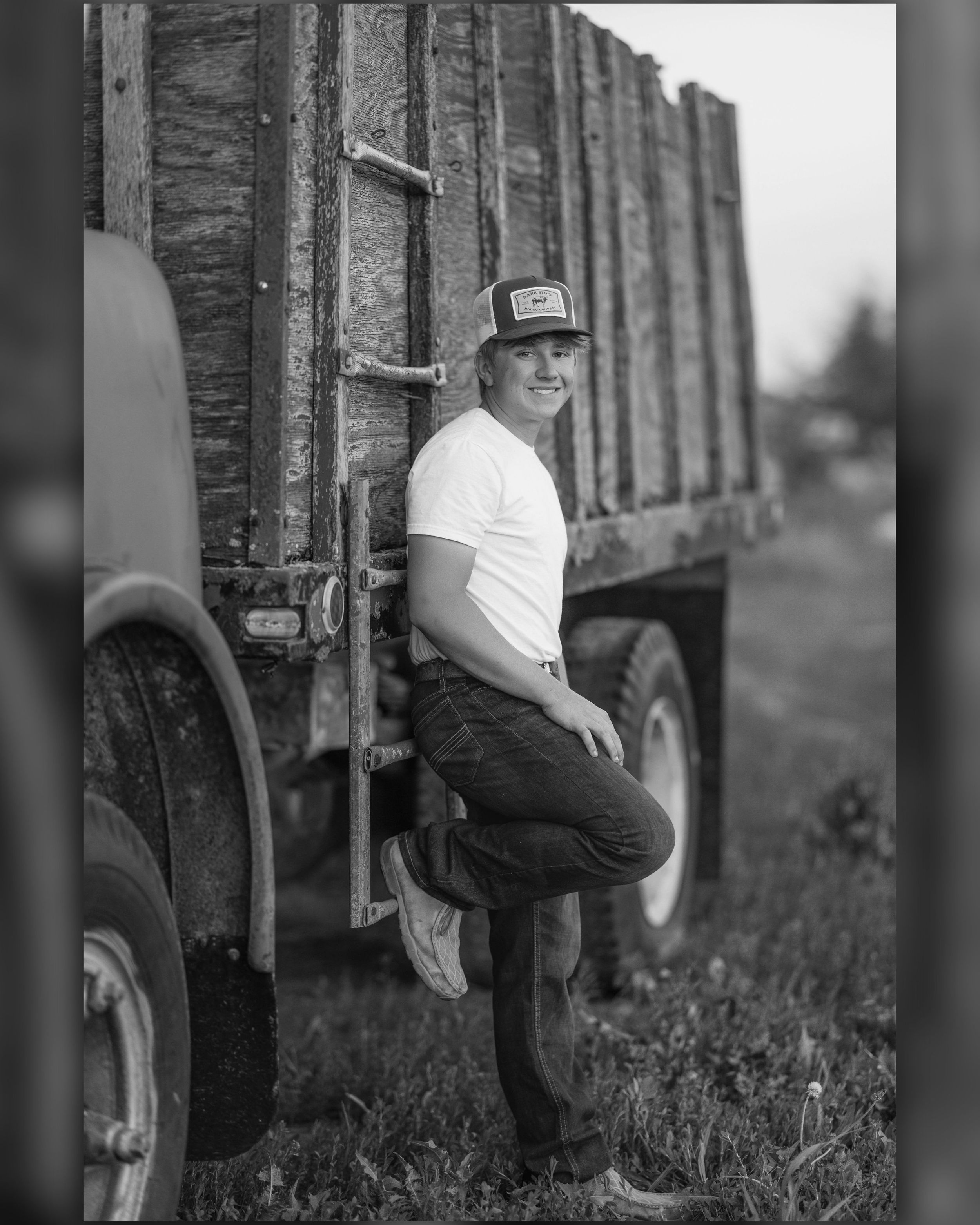 Young man in trucker hat and white t-shirt leaning against a farm truck in a black and white photo.