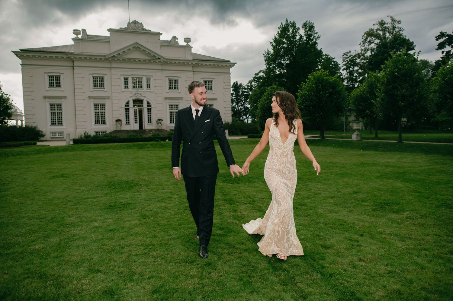 Bride and groom holding hands walking on green lawn before elegant white mansion on cloudy day.