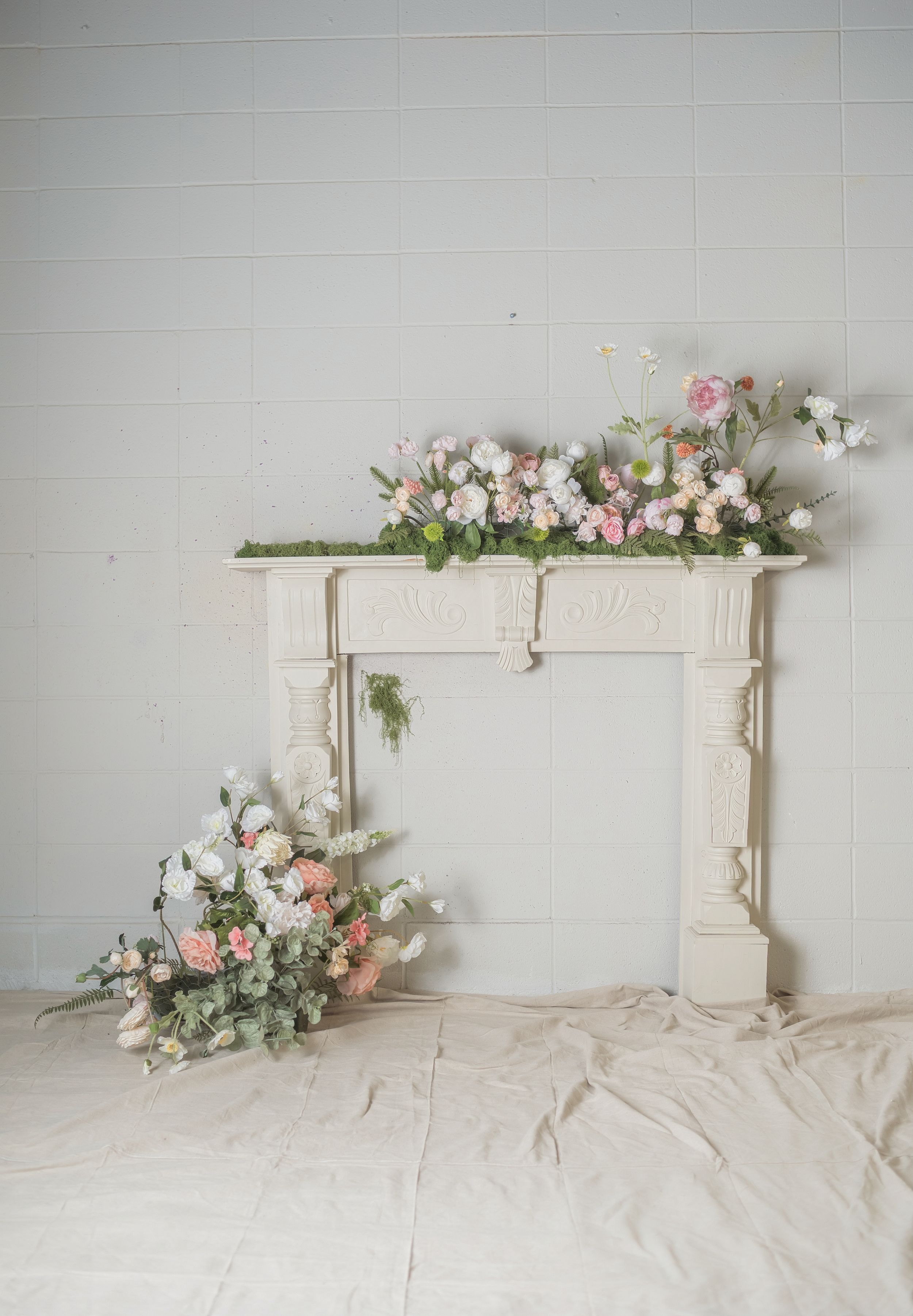White ornate fireplace mantel decorated with pink and white floral arrangements on top and floor beside it.