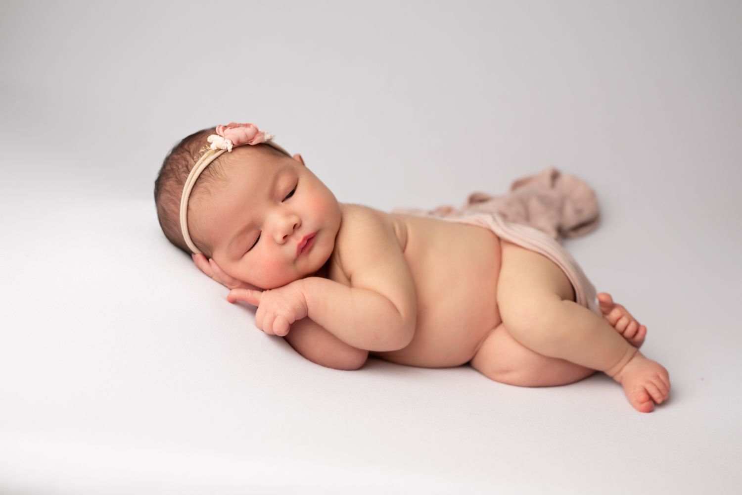 Peaceful newborn baby girl sleeping on white background wearing a delicate pink headband in a professional photo session.