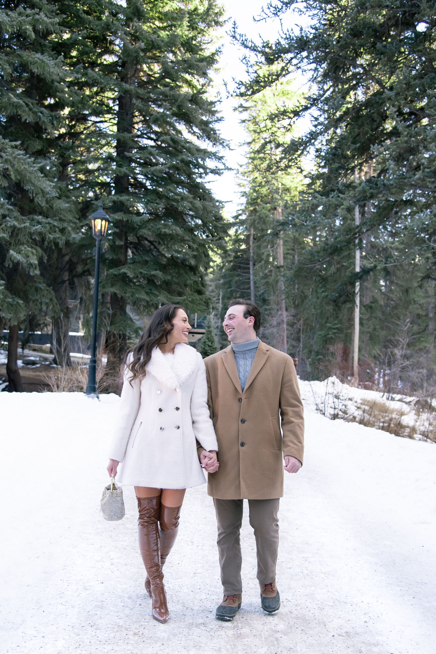 A couple walking hand in hand through a snowy forest path lined with evergreen trees on a winter day for Vail Colorado engagement photos