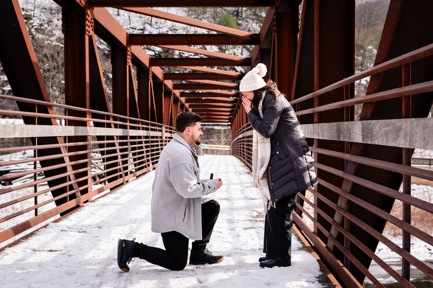 Man proposing on one knee to woman on snowy bridge in winter, romantic marriage proposal scene outdoors.