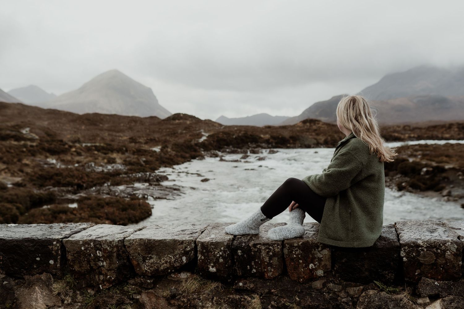 Woman in red dress stands on rocky terrain with snow-capped mountains and a white cottage in misty Scottish Highlands.